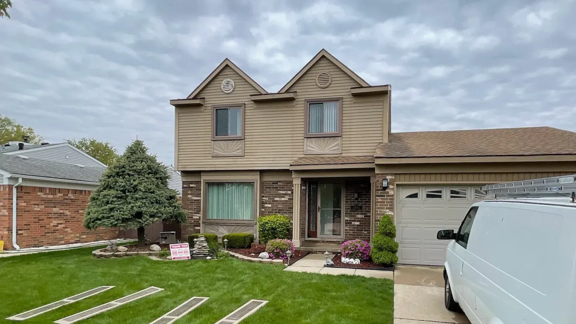 Two-story house with tan siding, brown brick, and a beige garage door under a cloudy sky.
