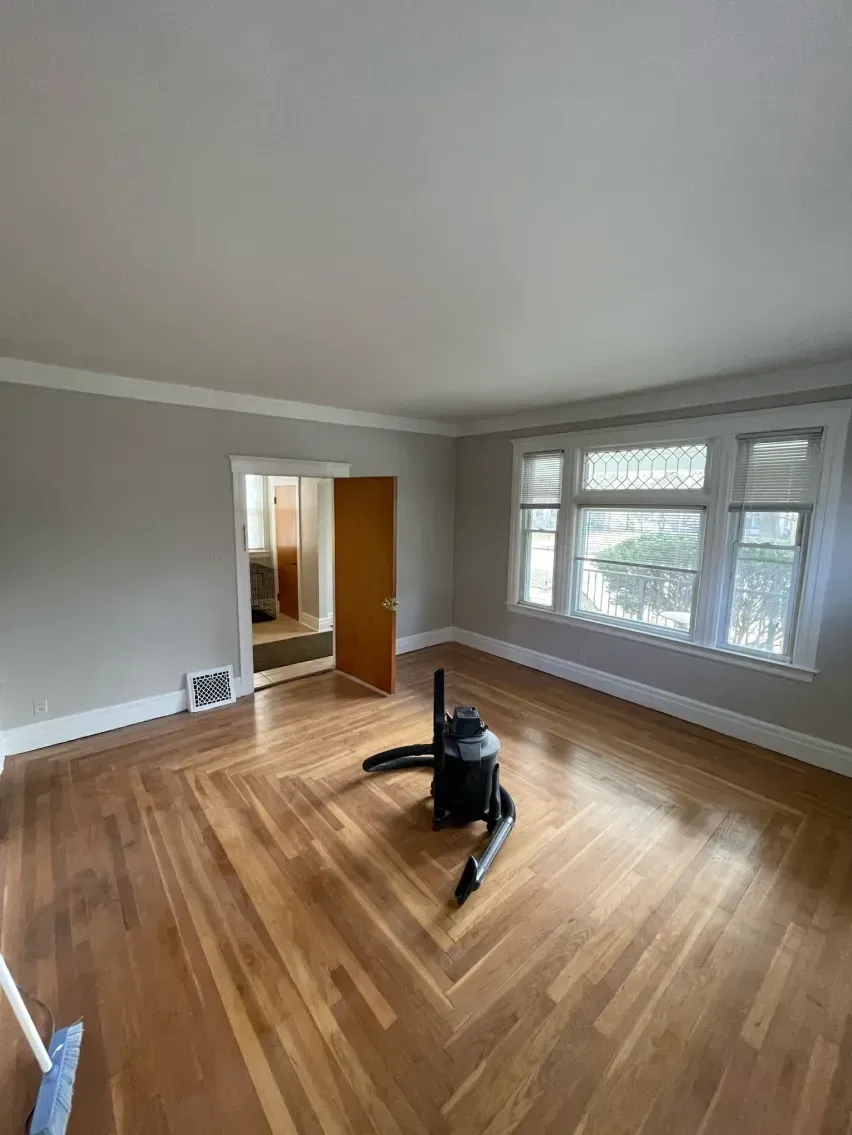 Empty room with parquet floor, gray walls, crown molding, and large windows. A vacuum cleaner sits in the middle.