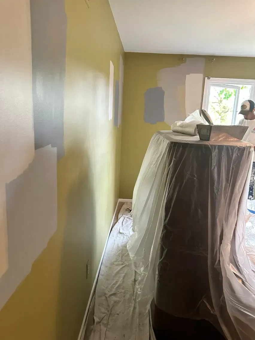 Interior wall with paint swatches in gray and yellow. Covered furniture and a window.