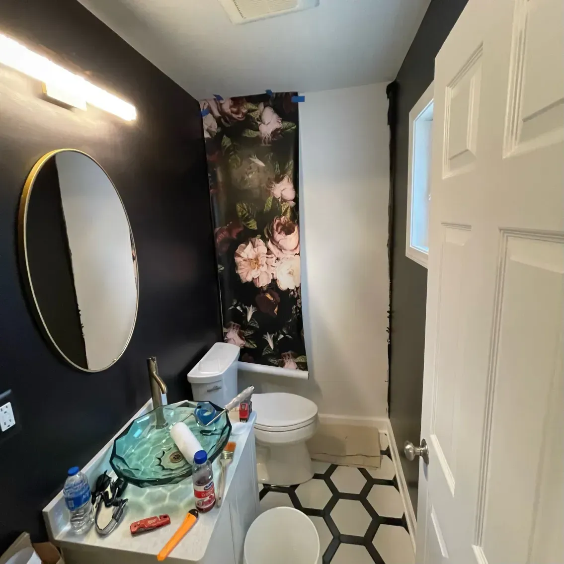 Small bathroom with dark blue wall, round mirror, floral shower curtain, hexagonal floor tiles, and white door.