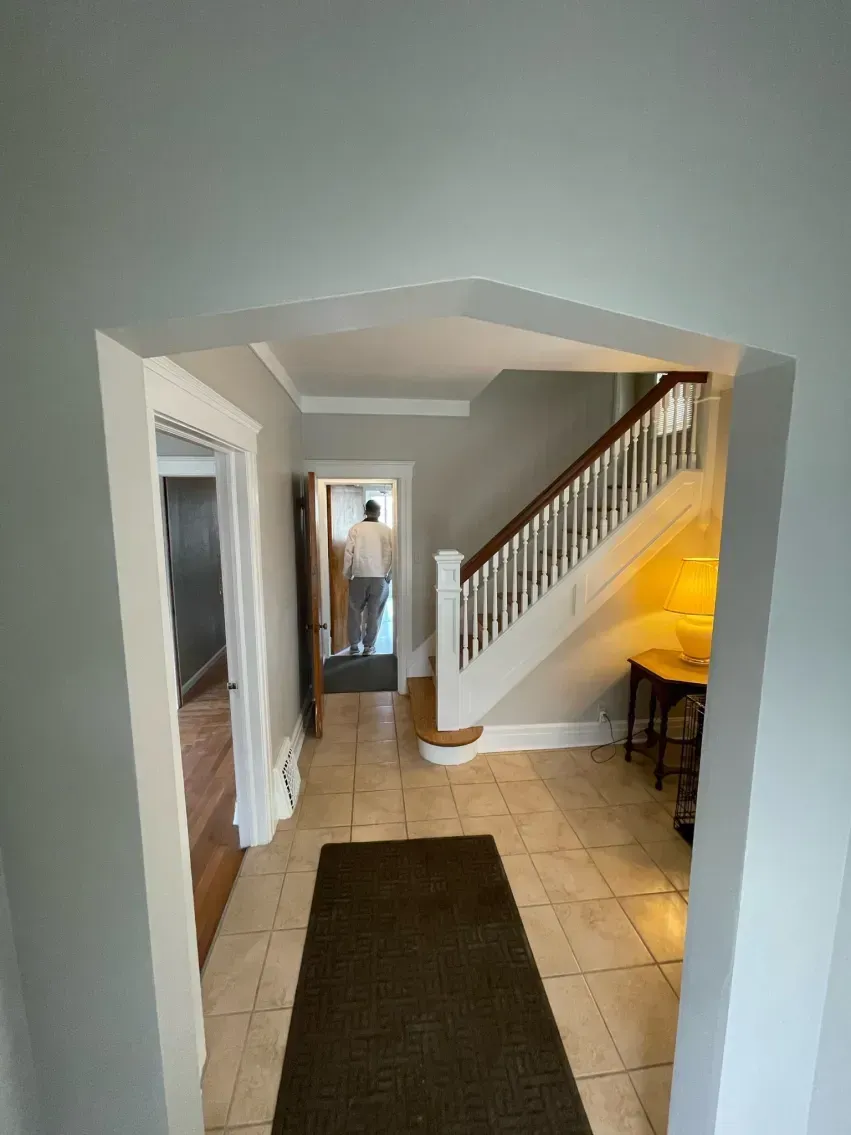 Hallway with staircase and person in distance; light tile floor, dark rug, light grey walls, white trim.