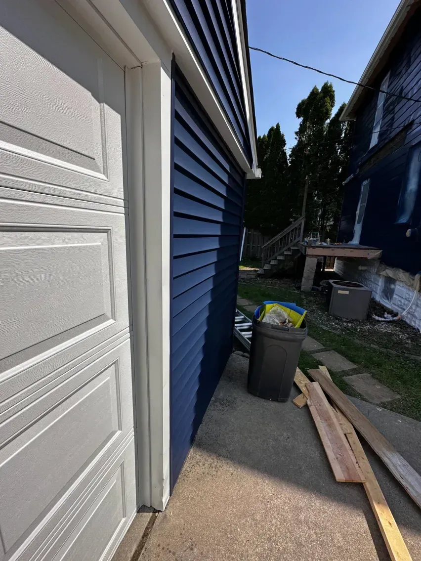 Garage with white door, blue siding, and trim. Piles of lumber and a trash can sit on the concrete.