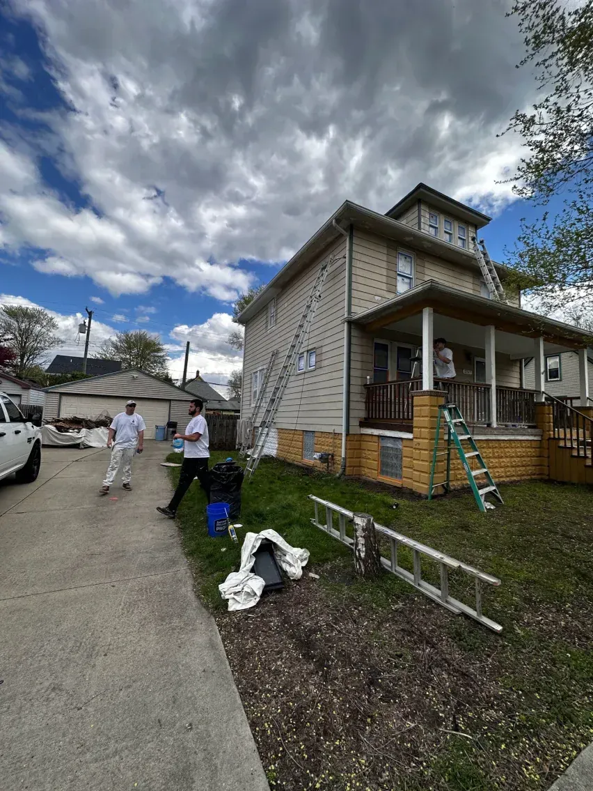 Two-story house being painted. Two painters on ladders. Cloudy sky. Driveway, grass.