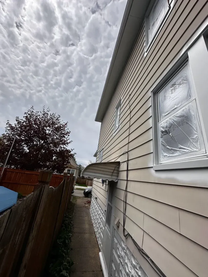 Side view of a two-story beige house, windows covered. A cloudy sky is overhead.