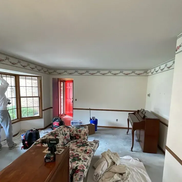 Living room renovation in progress: white walls, bay window, red door, floral sofa, piano.