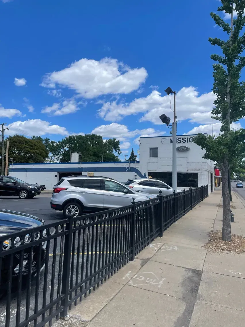 Cars parked next to a black fence in front of a white building under a cloudy blue sky.