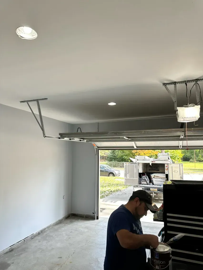 Man painting garage ceiling, open garage door with truck visible outside. Gray walls, recessed lights, and gray cement floor.