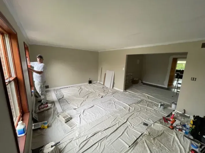 A person painting a room with beige walls and brown window frames. Drop cloths cover the floor.