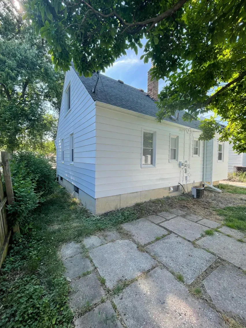 Side view of a white house with a blue-grey roof and a concrete patio. Trees frame the house.