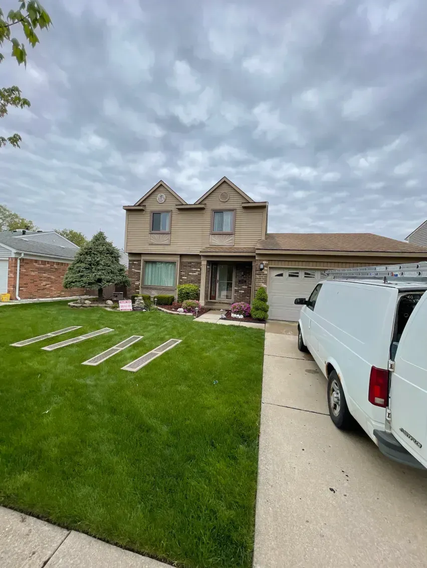Two-story house with tan stucco and stone facade, green lawn, and white van parked in driveway.