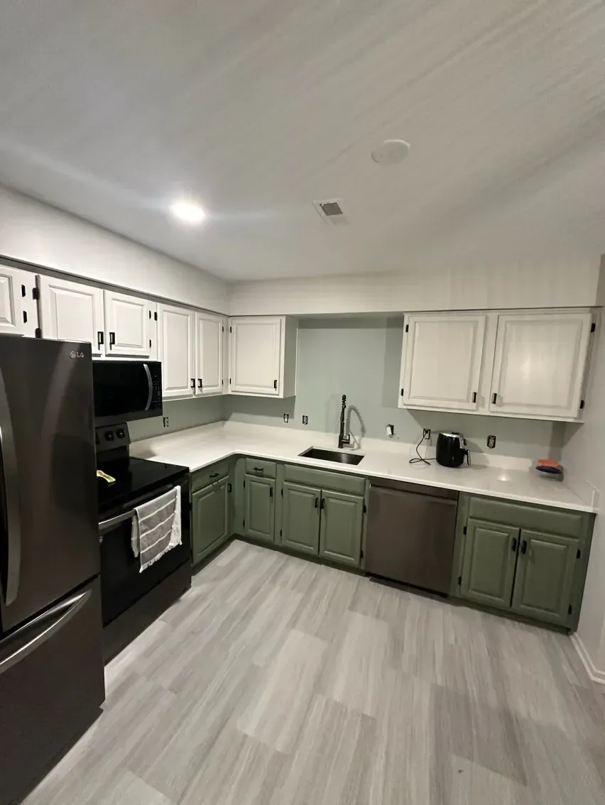 L-shaped kitchen with white upper cabinets and green lower cabinets, stainless steel appliances, and gray flooring.