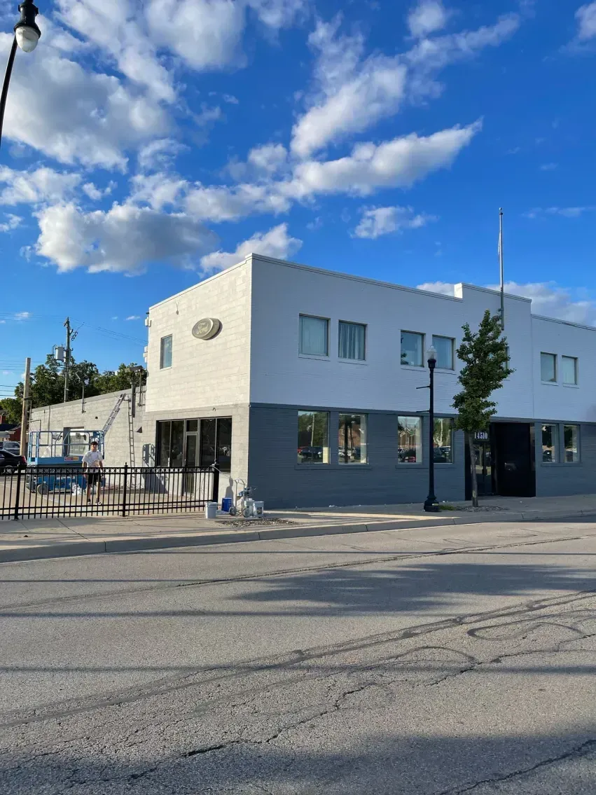 Two-story white and gray building with large windows, street view, blue sky with clouds.