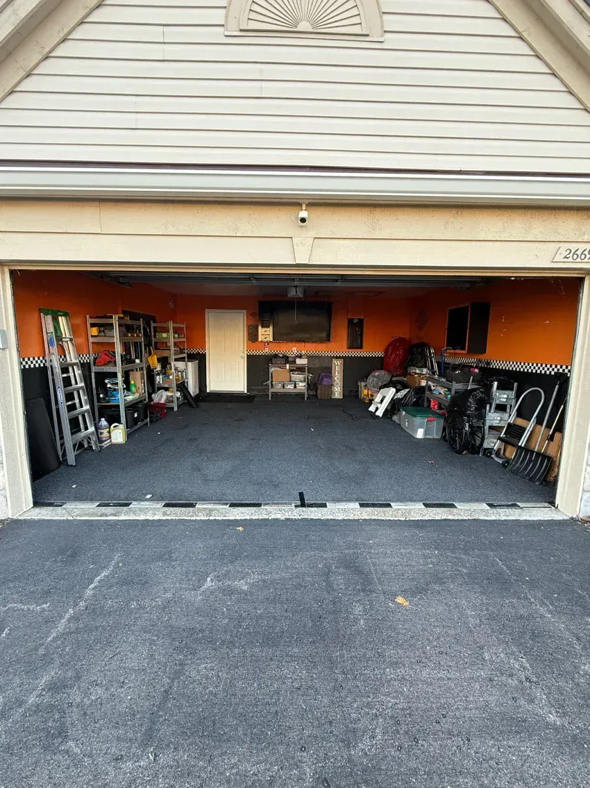 Open garage with dark flooring, orange walls, and storage. Piles of items line walls. Asphalt driveway.