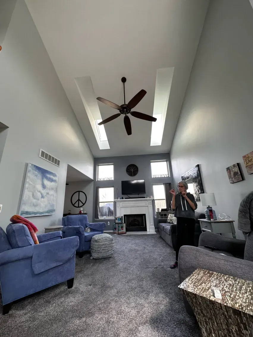 Living room with tall ceiling, blue furniture, fireplace, and a ceiling fan; person taking a picture.