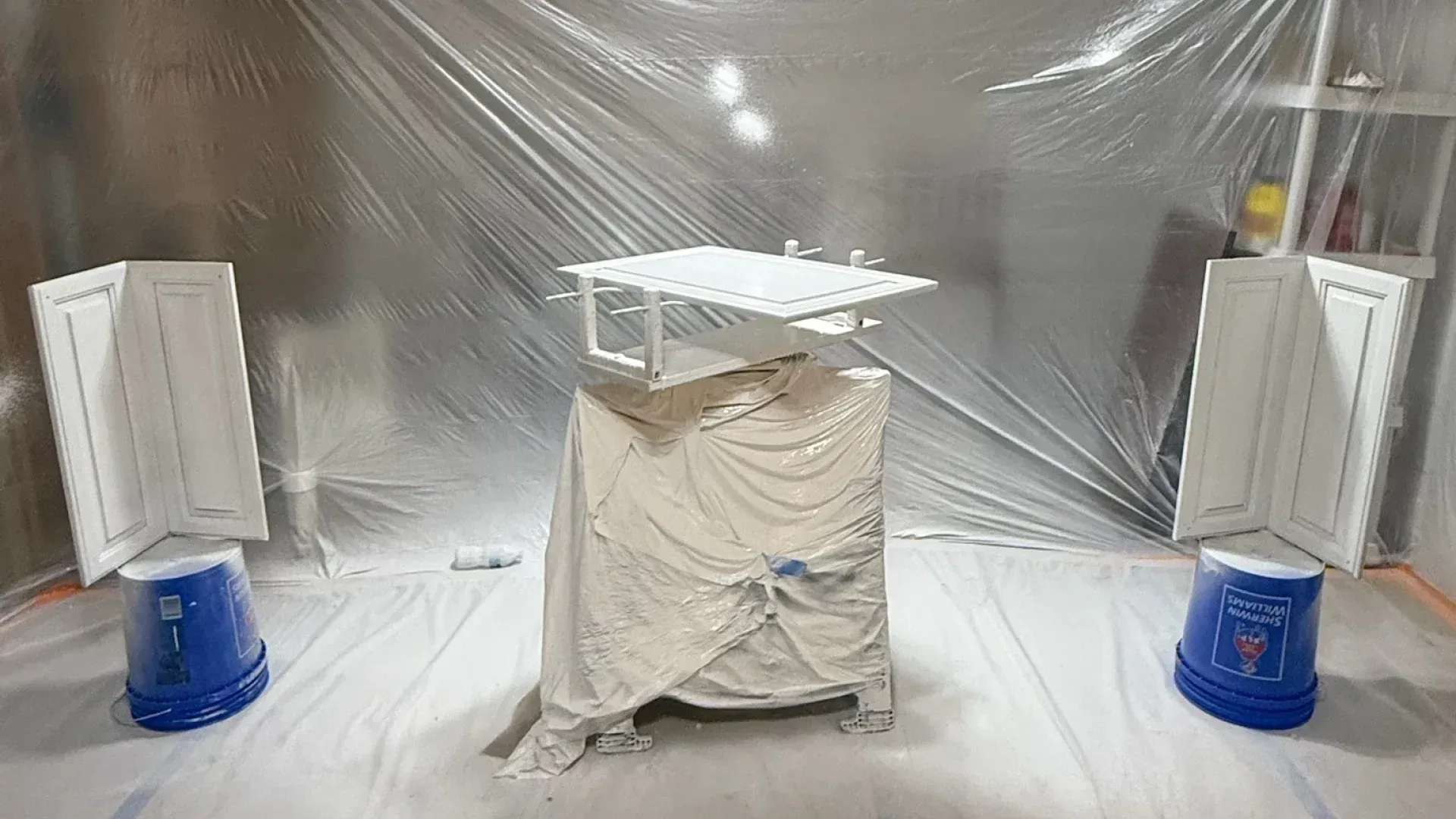 White cabinets and drawers covered for painting in a room draped with plastic.
