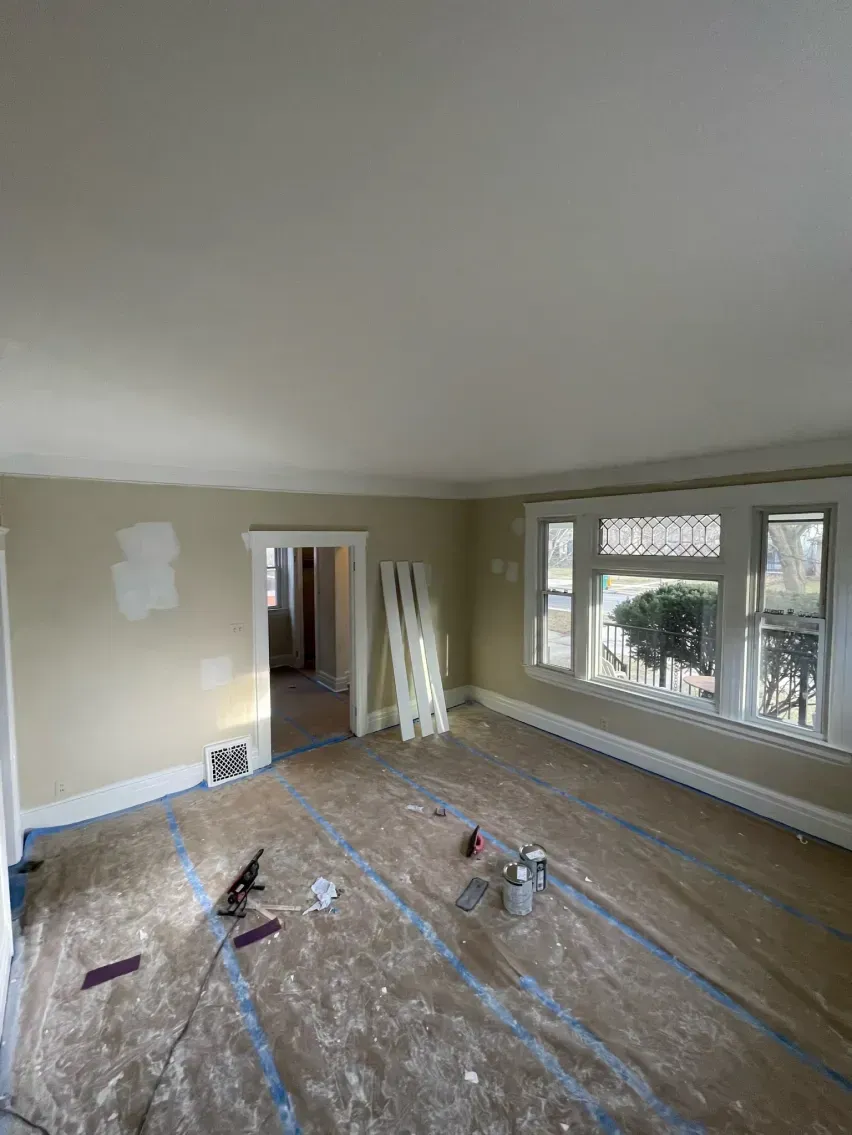 Living room renovation in progress. Beige walls, wood floor covered in blue tape, and window with outdoor view.