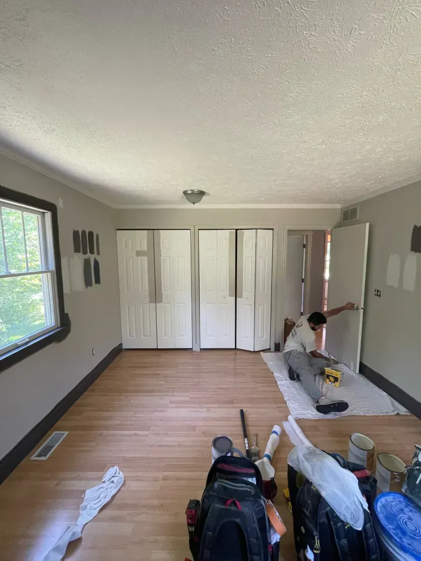 Person painting door in a room with light wood floors, white doors, and grey walls.