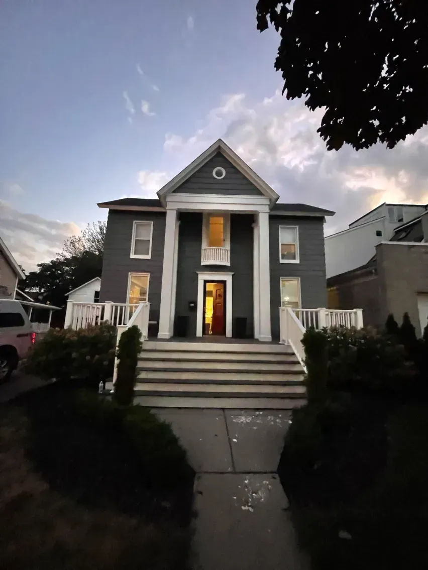 Gray house with white columns, steps leading to the front door, bushes and a cloudy sky.