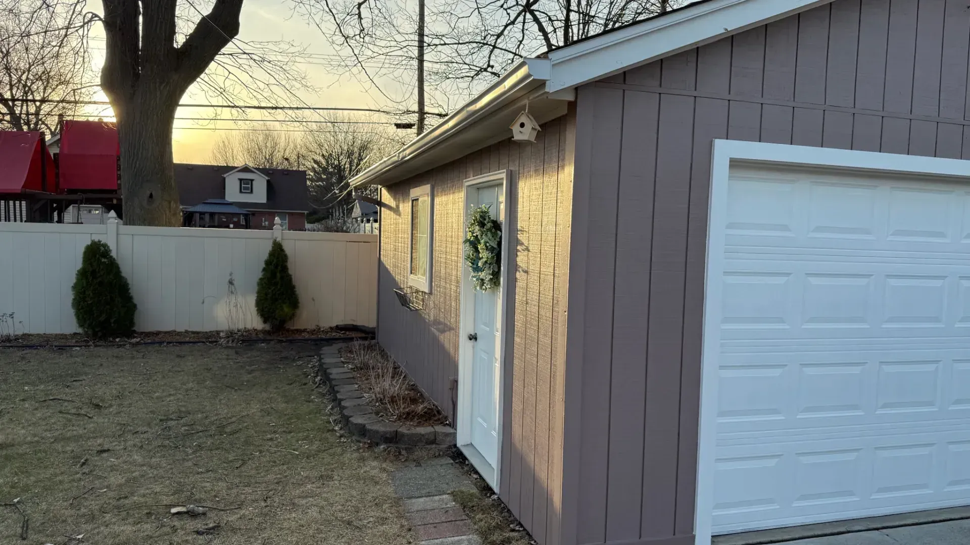 Garage with white door and trim, purple siding, wreath. Backyard setting, fence, small trees.