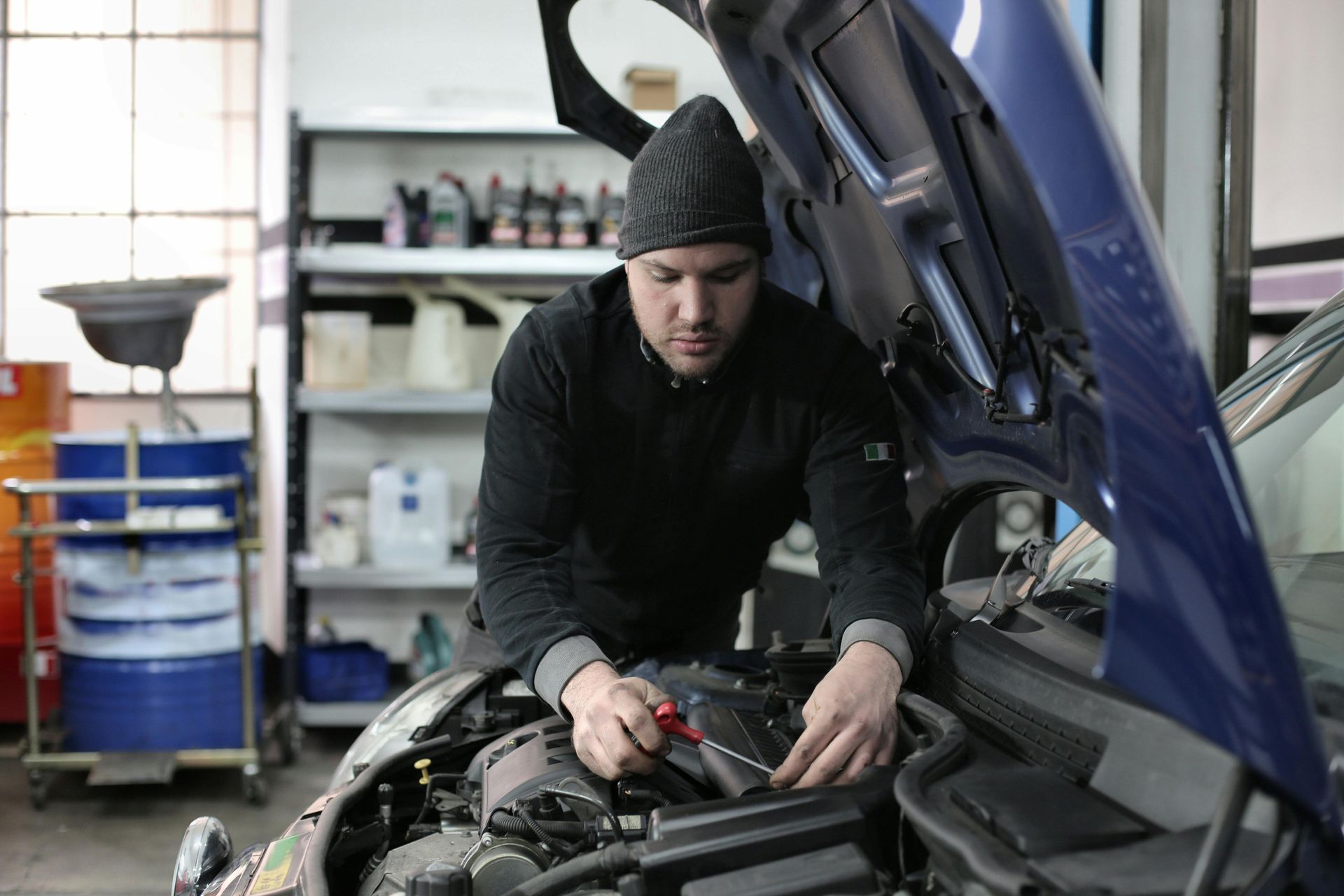 Mechanic in a beanie works on a blue car engine in a garage, using a screwdriver.