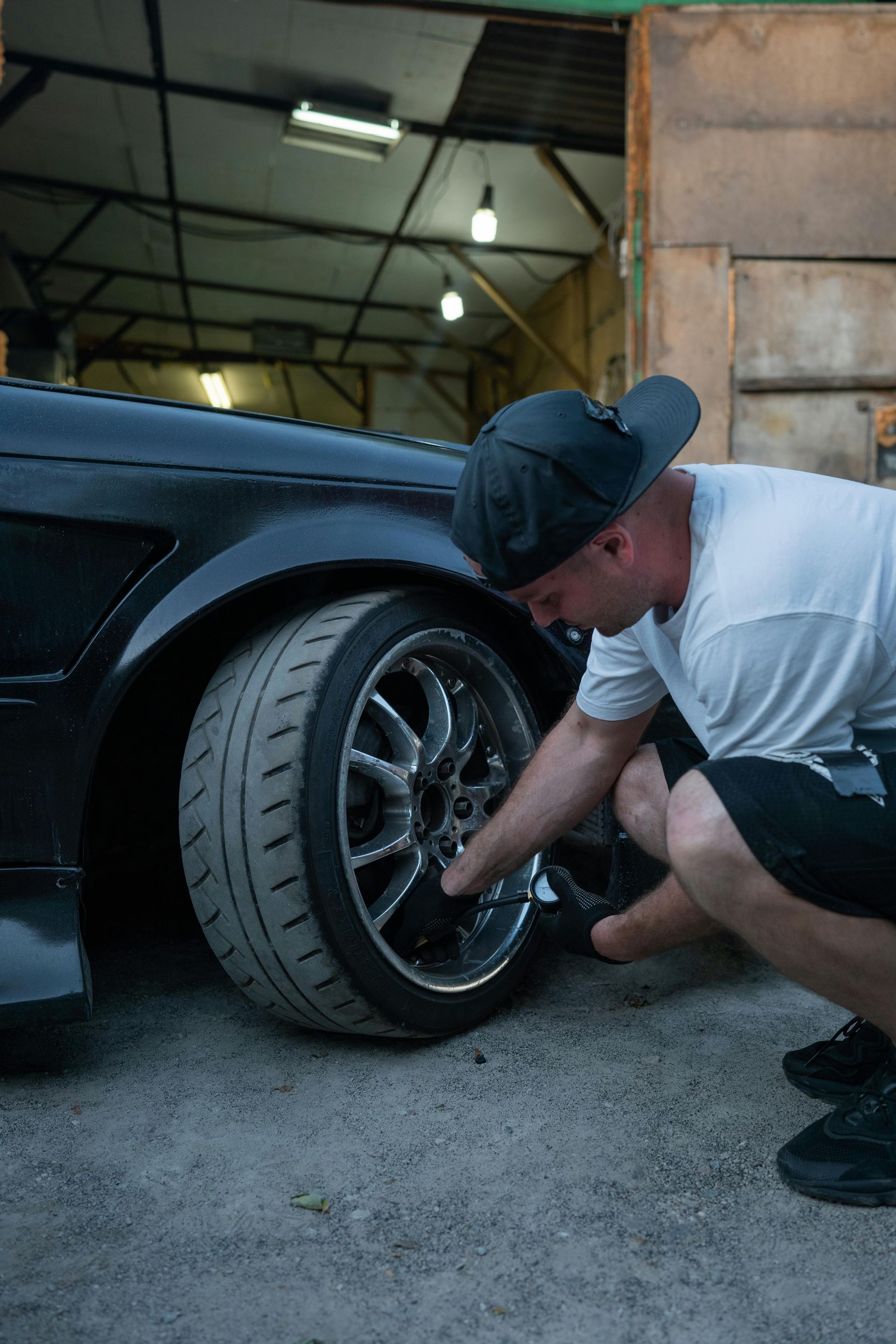 Man in cap inspecting car wheel, likely in a garage setting. Black car, chrome rim, and worn tires.
