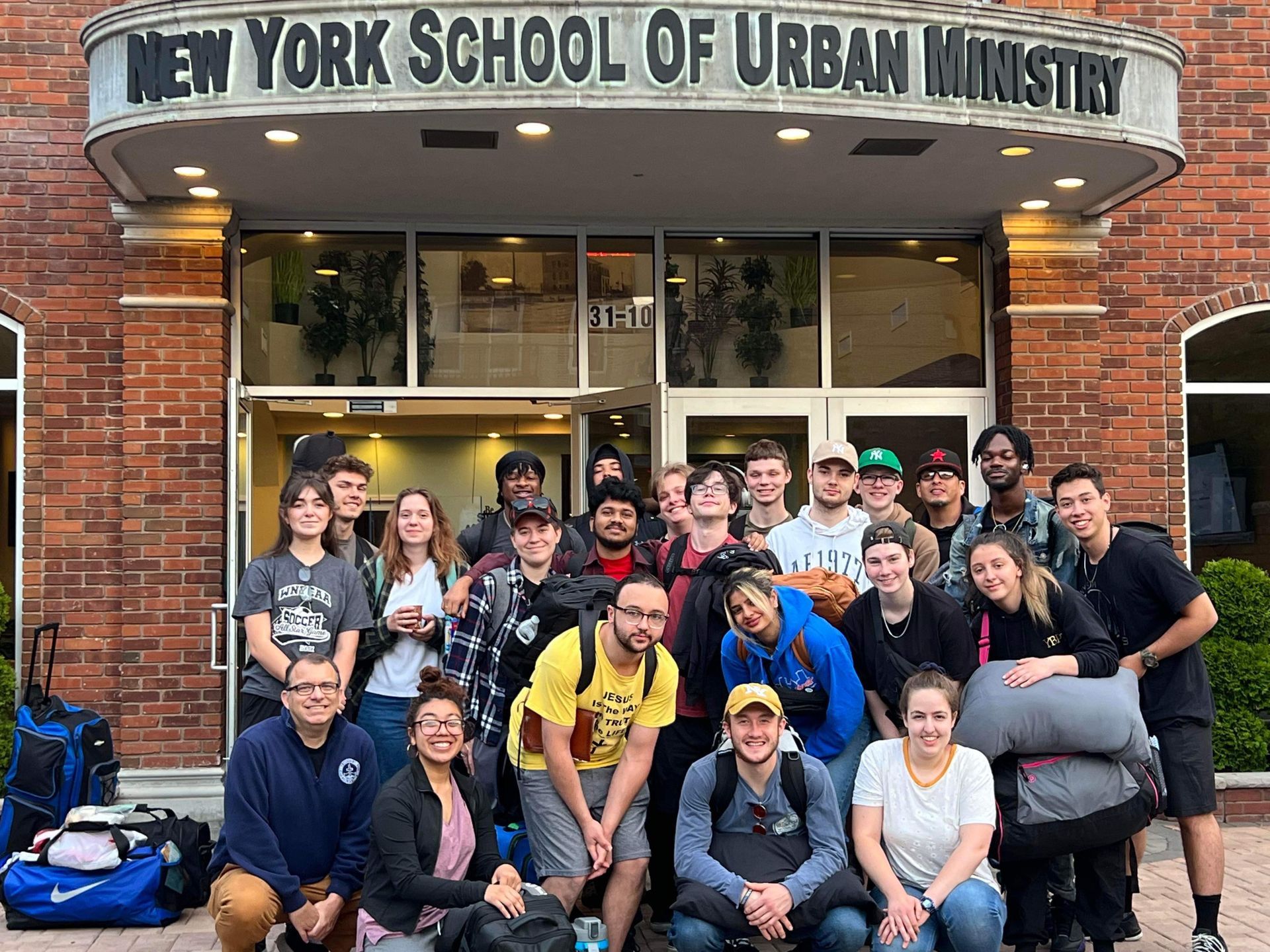 Group of young people posing outside the New York School of Urban Ministry. They are smiling and carrying luggage.