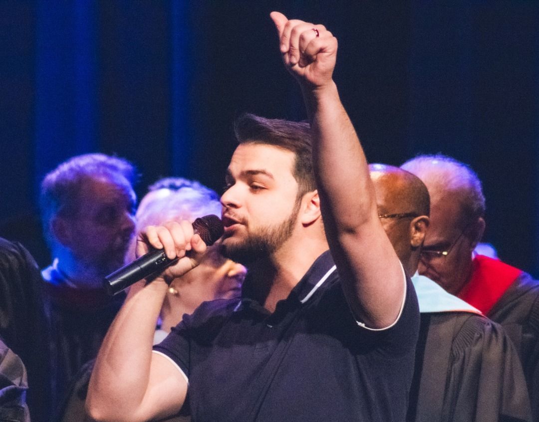 Man with a microphone singing on stage, raising his right arm. Other people are blurred in the background.