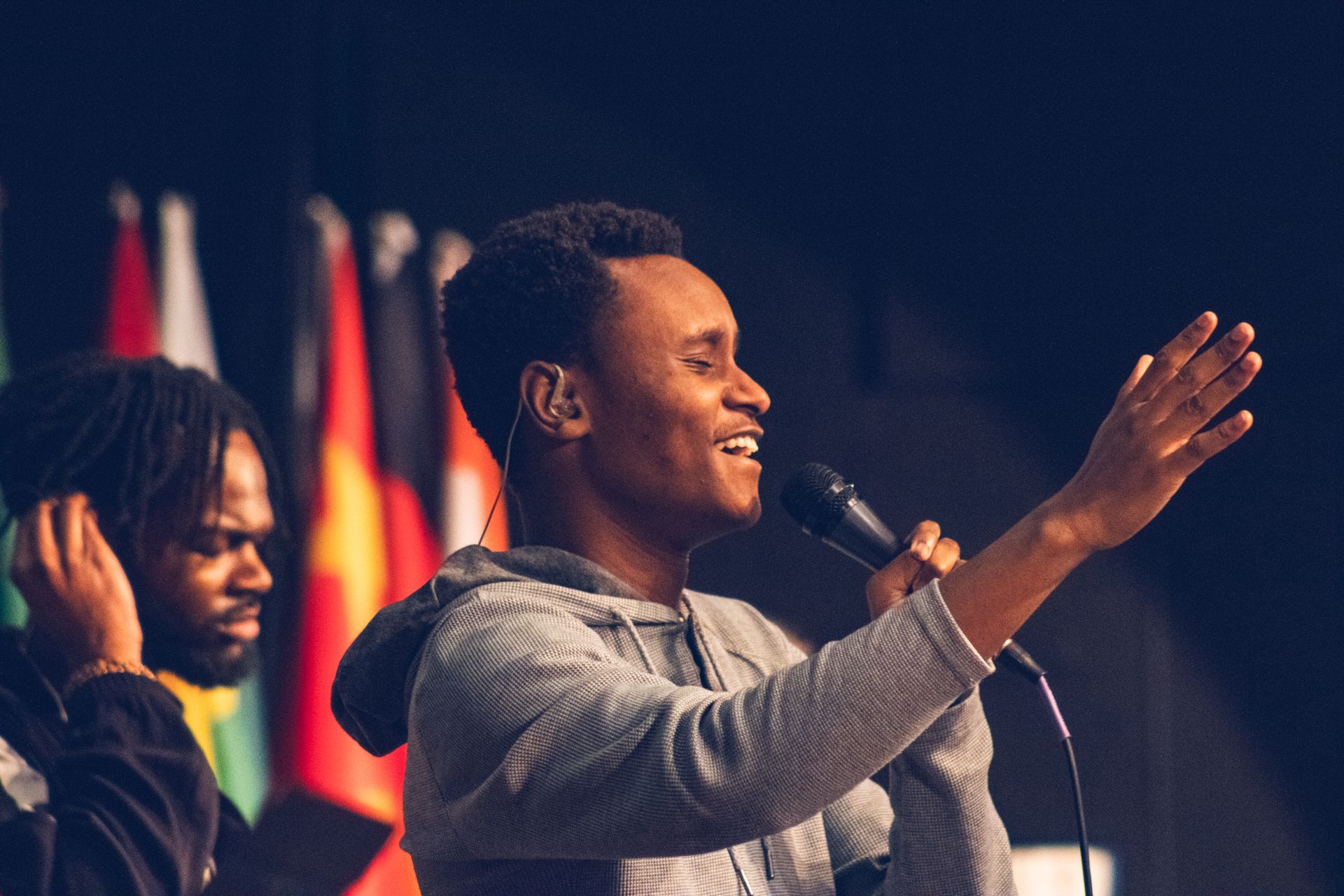 Two Black men on a stage; one singing into a microphone with his arm raised, smiling; the other to his side listening. 