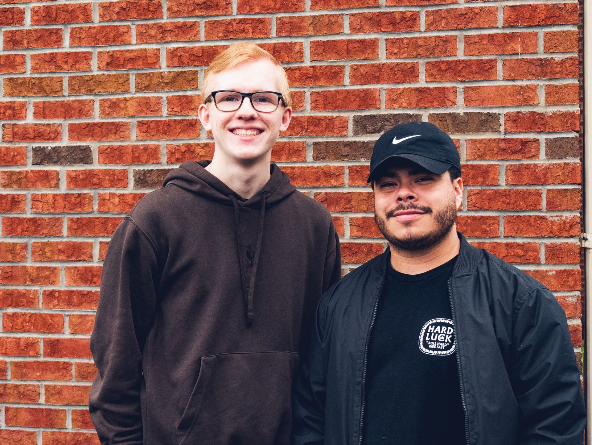 Two young men stand in front of a red brick wall. 
