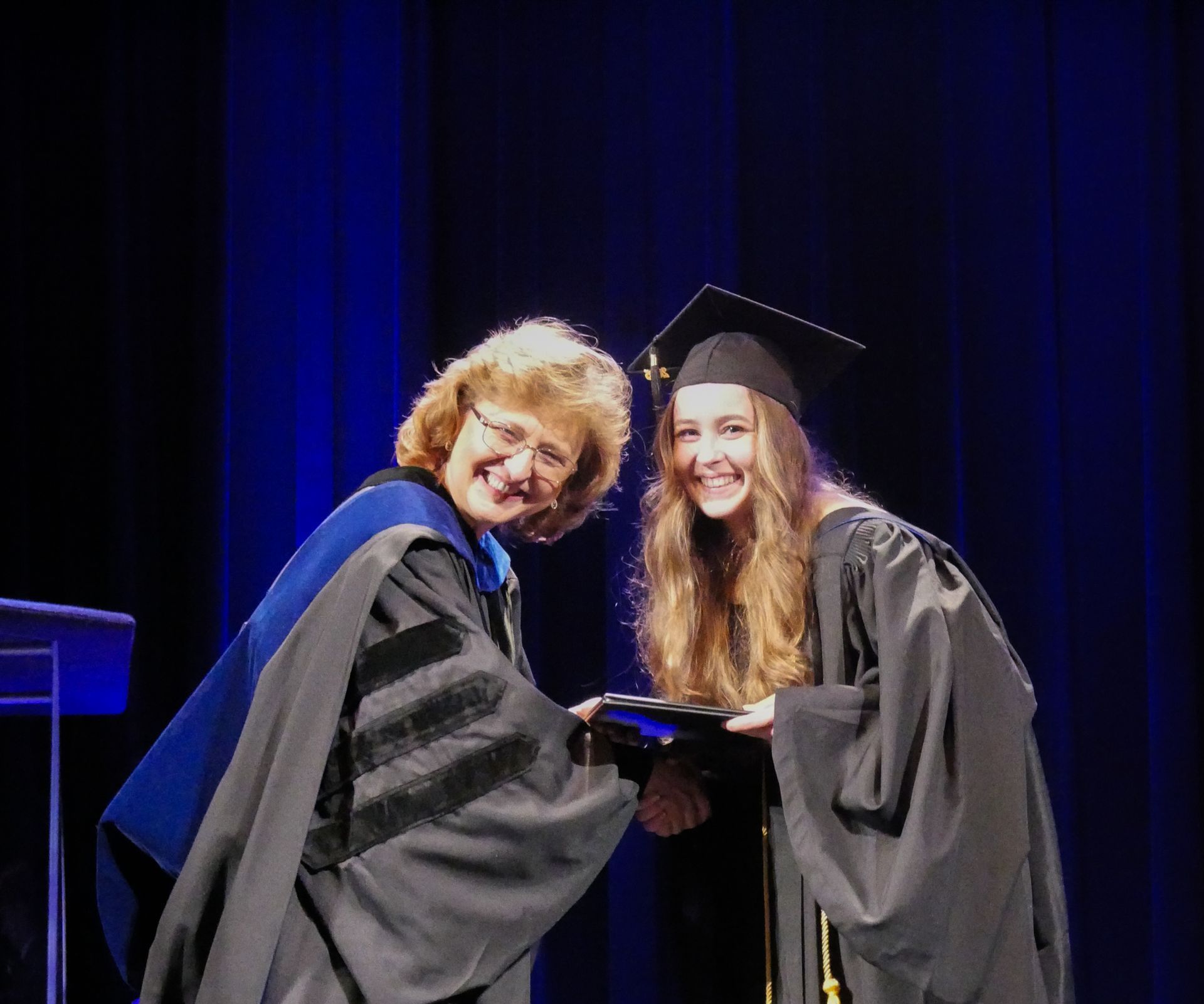 Woman in academic regalia presents a diploma to a graduating student on a stage with a blue backdrop; both smile.
