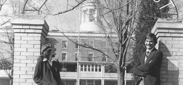 A man and a woman pose outside a brick building. The man has his arms crossed and the woman is wearing a coat.