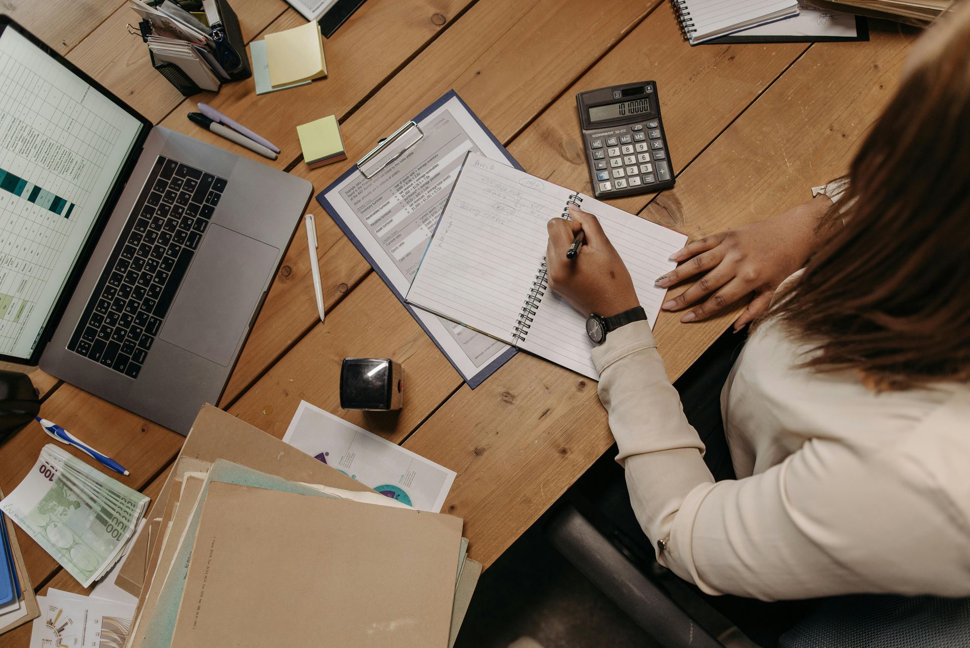 Woman writing in a notebook at a wooden desk, surrounded by a laptop, documents, calculator, and office supplies.
