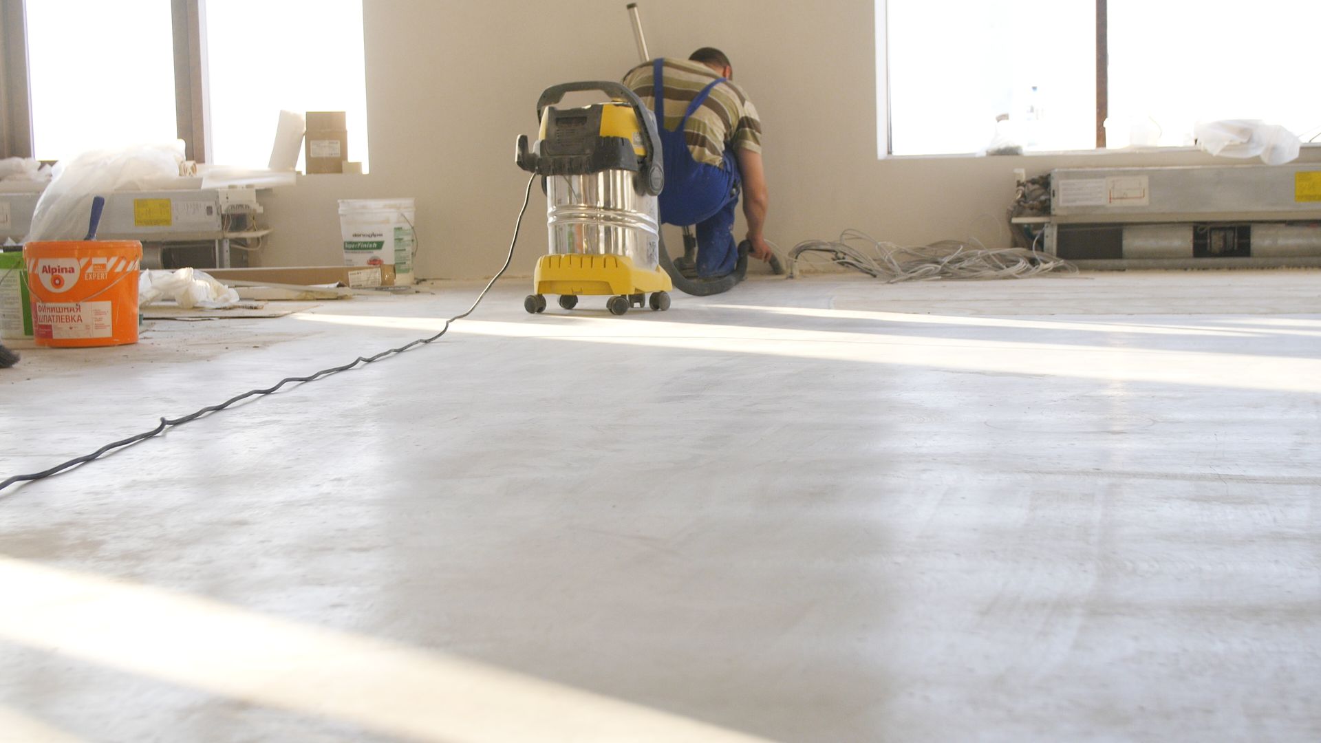 Person vacuuming a light-colored floor in a room under construction, with buckets and materials nearby.