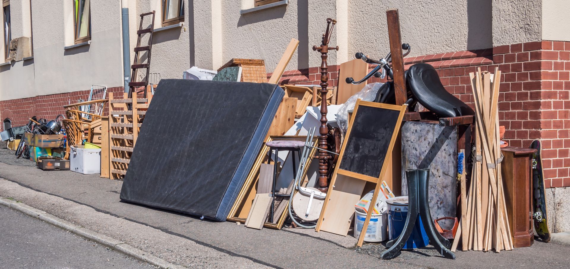 Pile of discarded furniture and household items on a sidewalk near a building with brick accents.