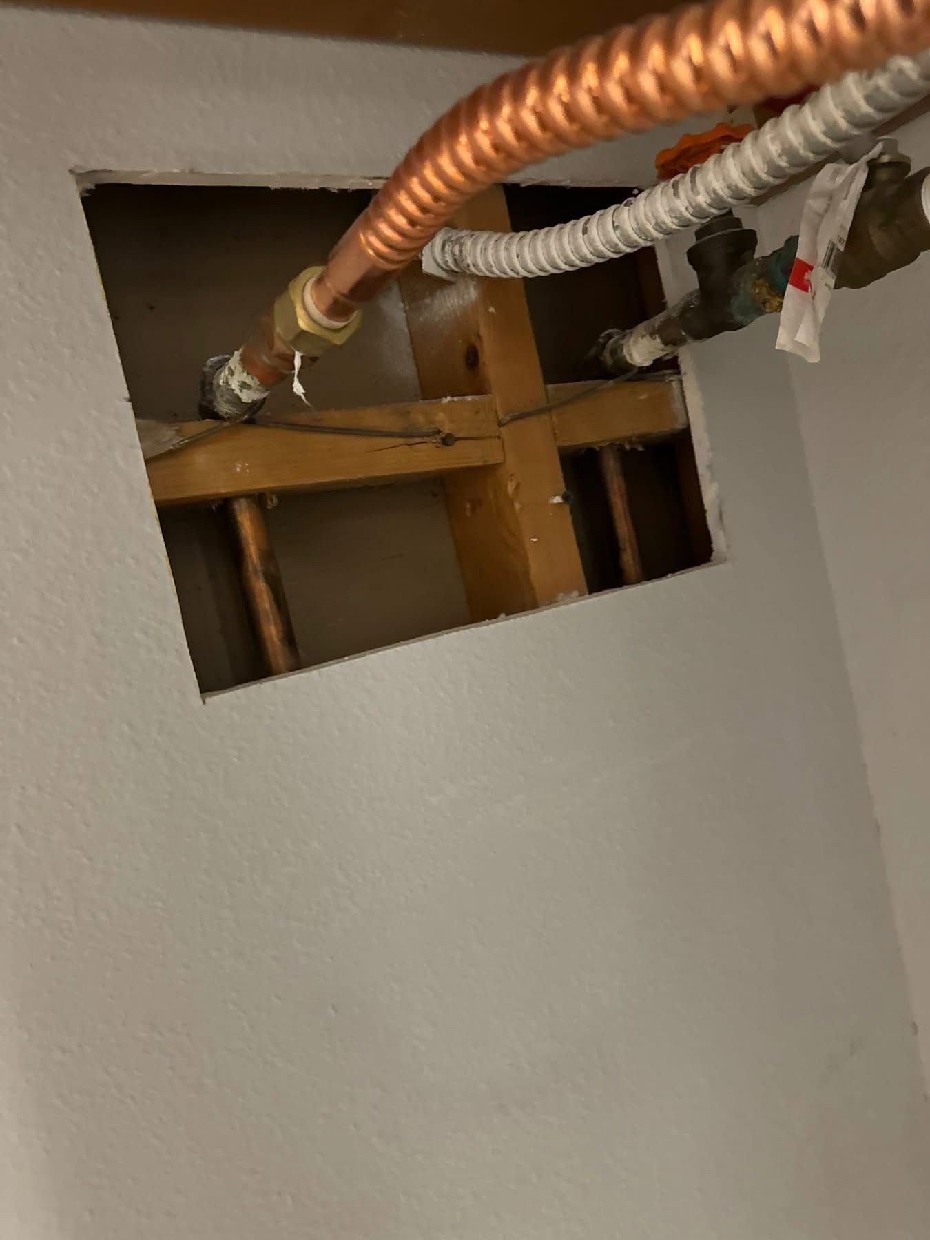 Square hole in wall exposing copper pipes, wooden beams, and a white plastic pipe.