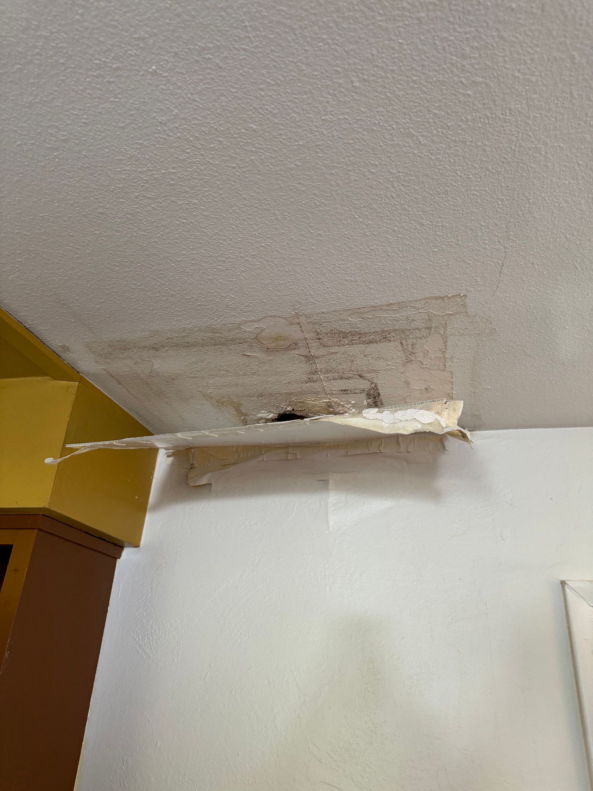 Water damaged ceiling with peeling paint, near a yellow cabinet and white wall.