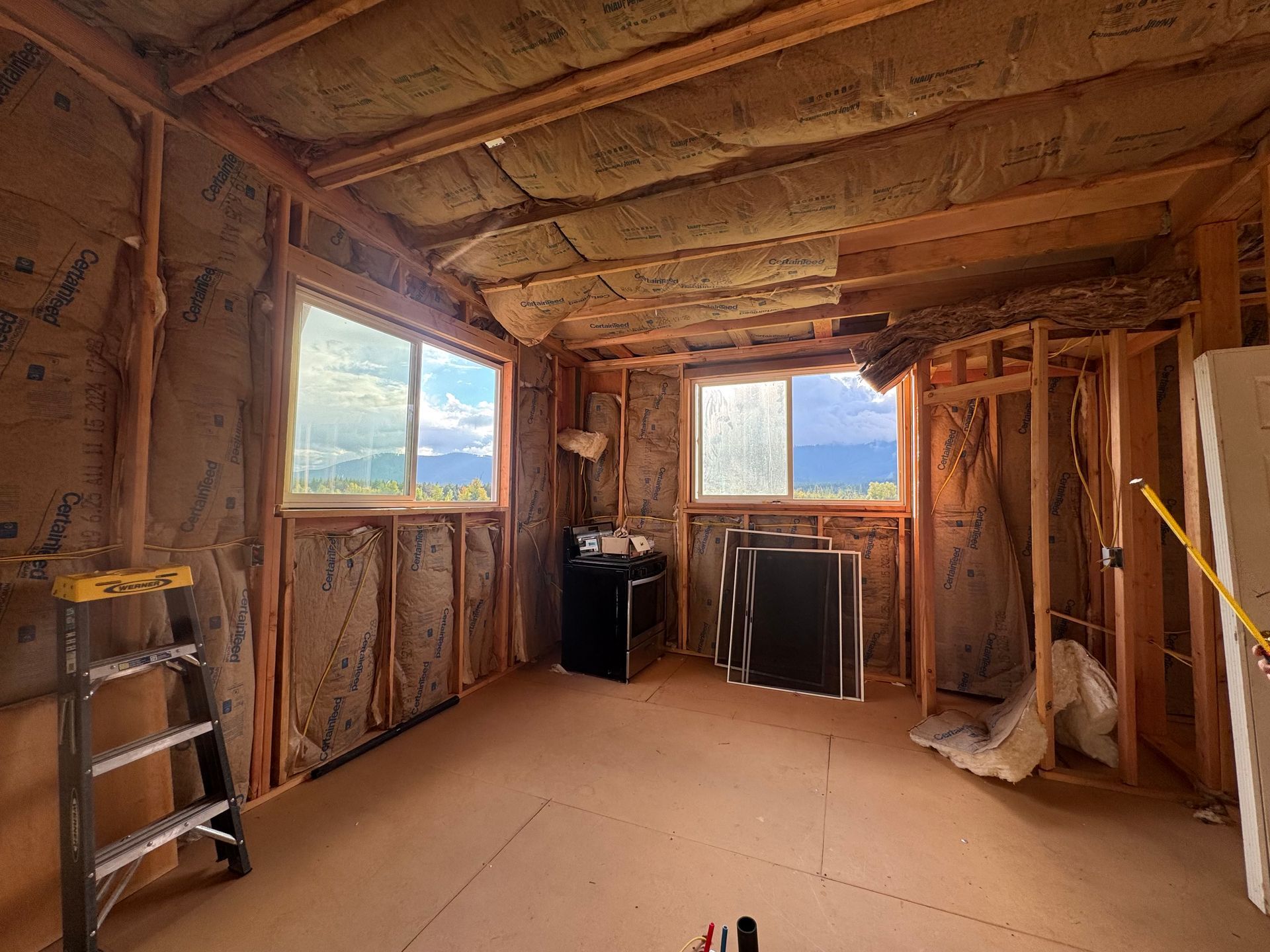 Interior of a room under construction, with wooden framing, insulation, windows, and a ladder.