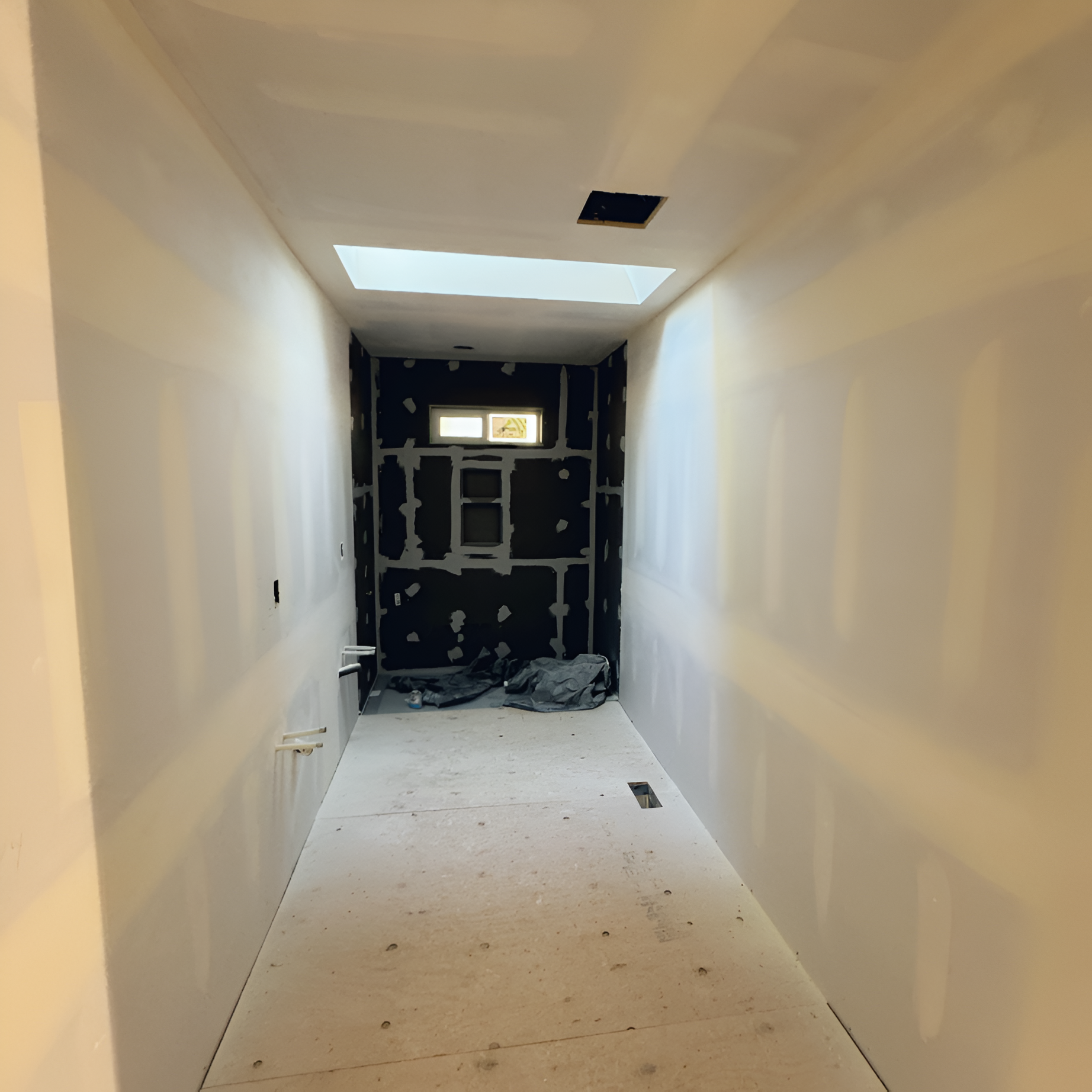 Narrow hallway under construction; drywall, black tile, skylight, and unfinished floor.