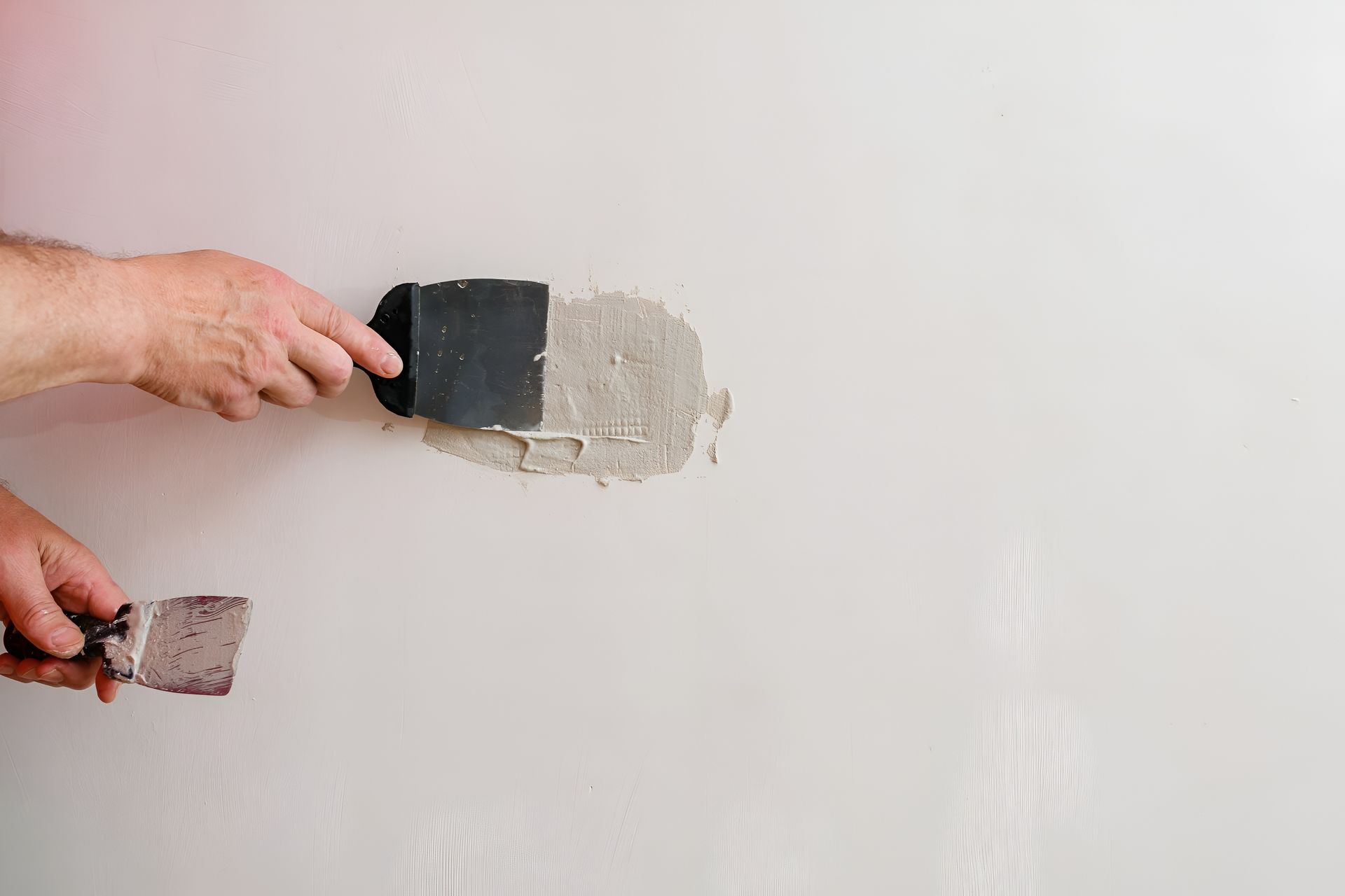 Hands using a putty knife to apply spackle to a wall, filling a damaged area.