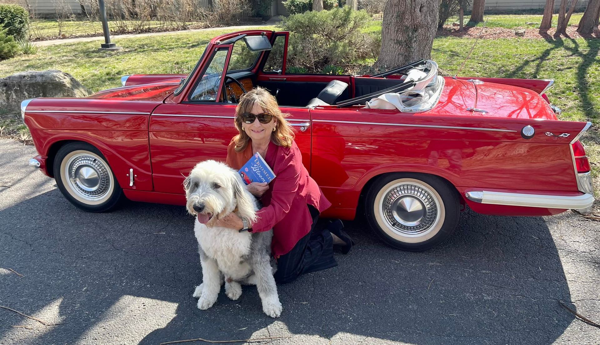 A woman, Susan Armstrong, is kneeling down next to a dog in front of a red car.