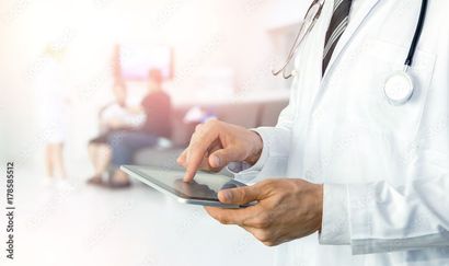 Doctor in white coat using tablet, stethoscope, patients in background.