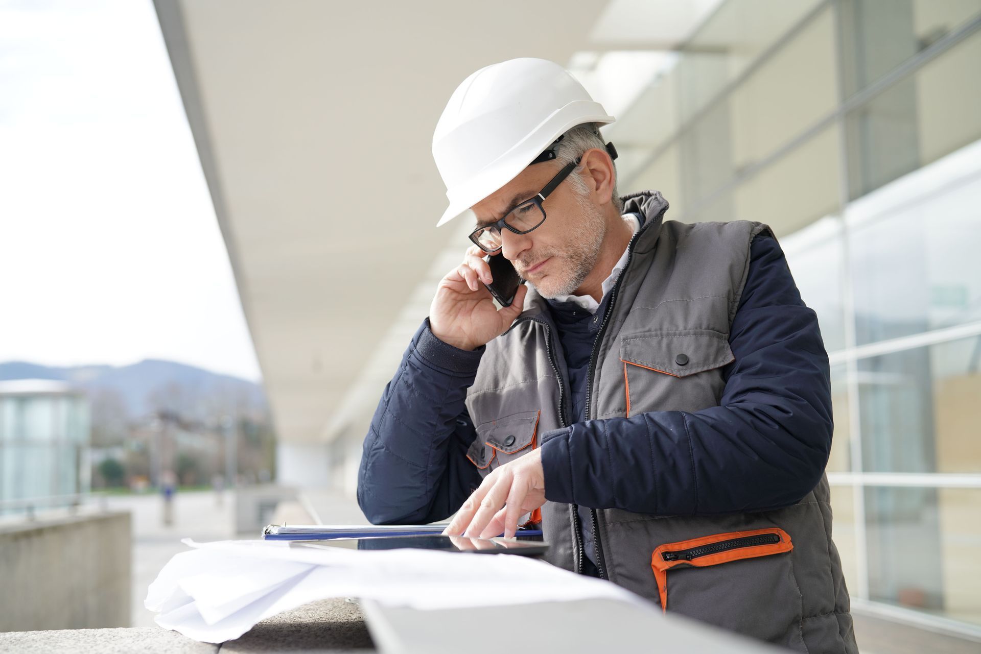 Construction worker in a hard hat, jacket, and glasses, on the phone, looking at blueprints outside a building.