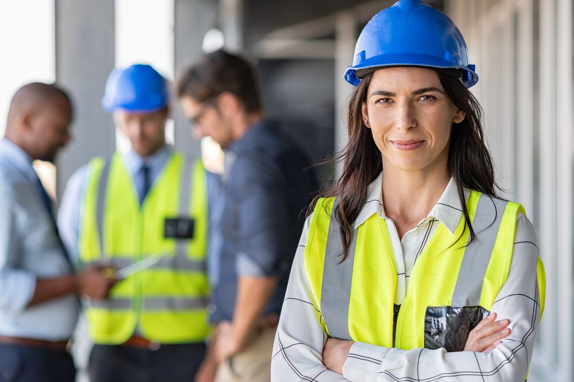 Woman in hard hat and safety vest, arms crossed, smiling. Construction site with colleagues blurred in background.