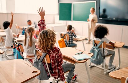 Children raising hands in a classroom, teacher at the whiteboard, desks arranged.