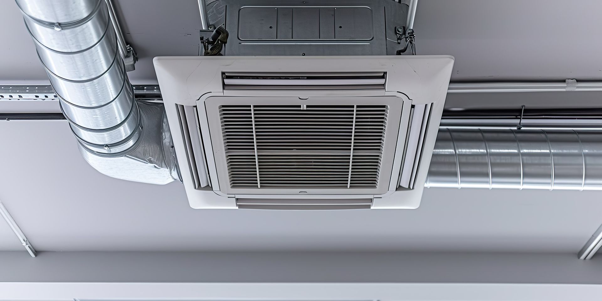 Ceiling-mounted air conditioning unit with metal ductwork, set against a white ceiling.