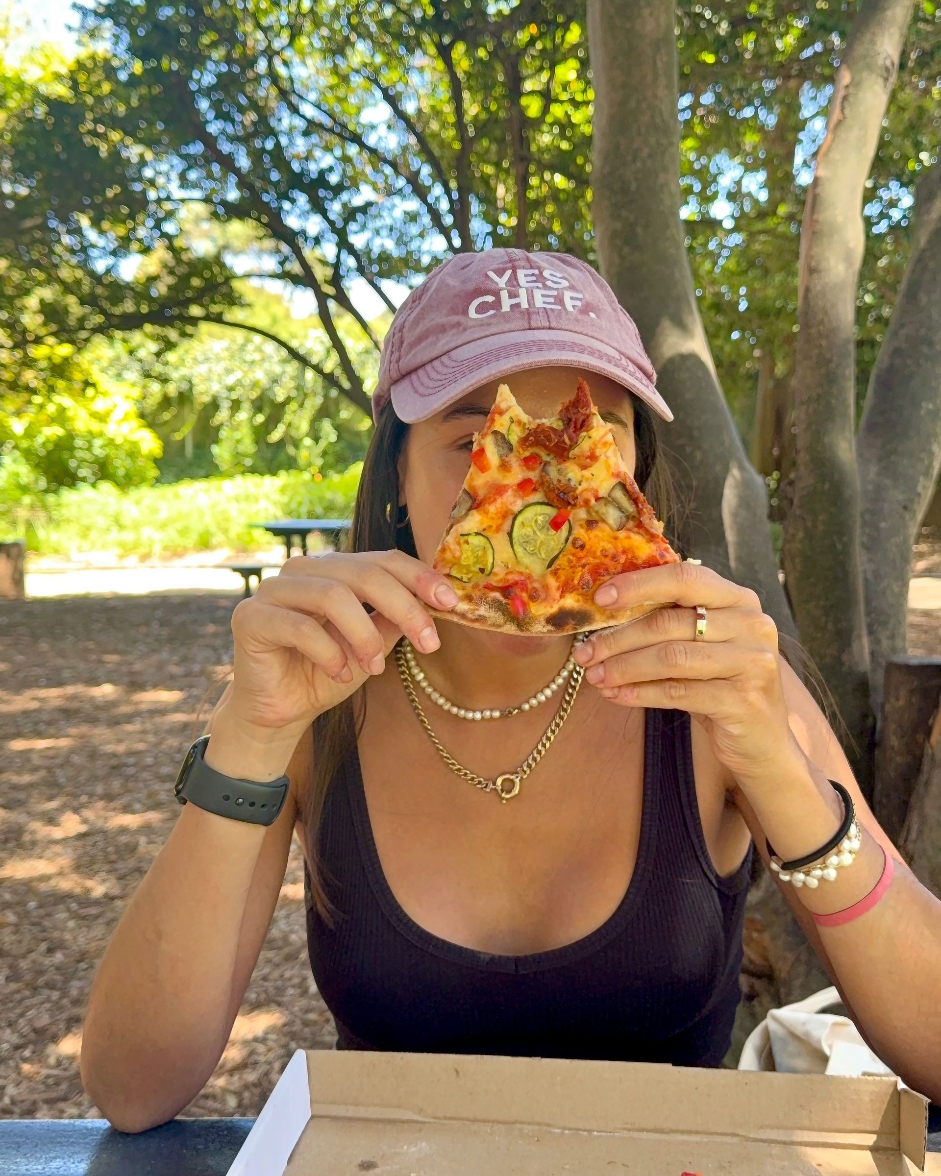 Woman holding pizza slice in front of her face, outdoors under a tree. 