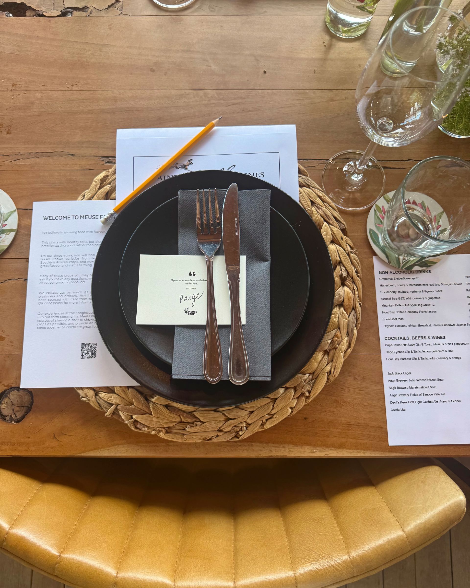 Place setting on a wooden table. Black plate, silverware, name card, menu, pencil, water glass, wicker placemat.