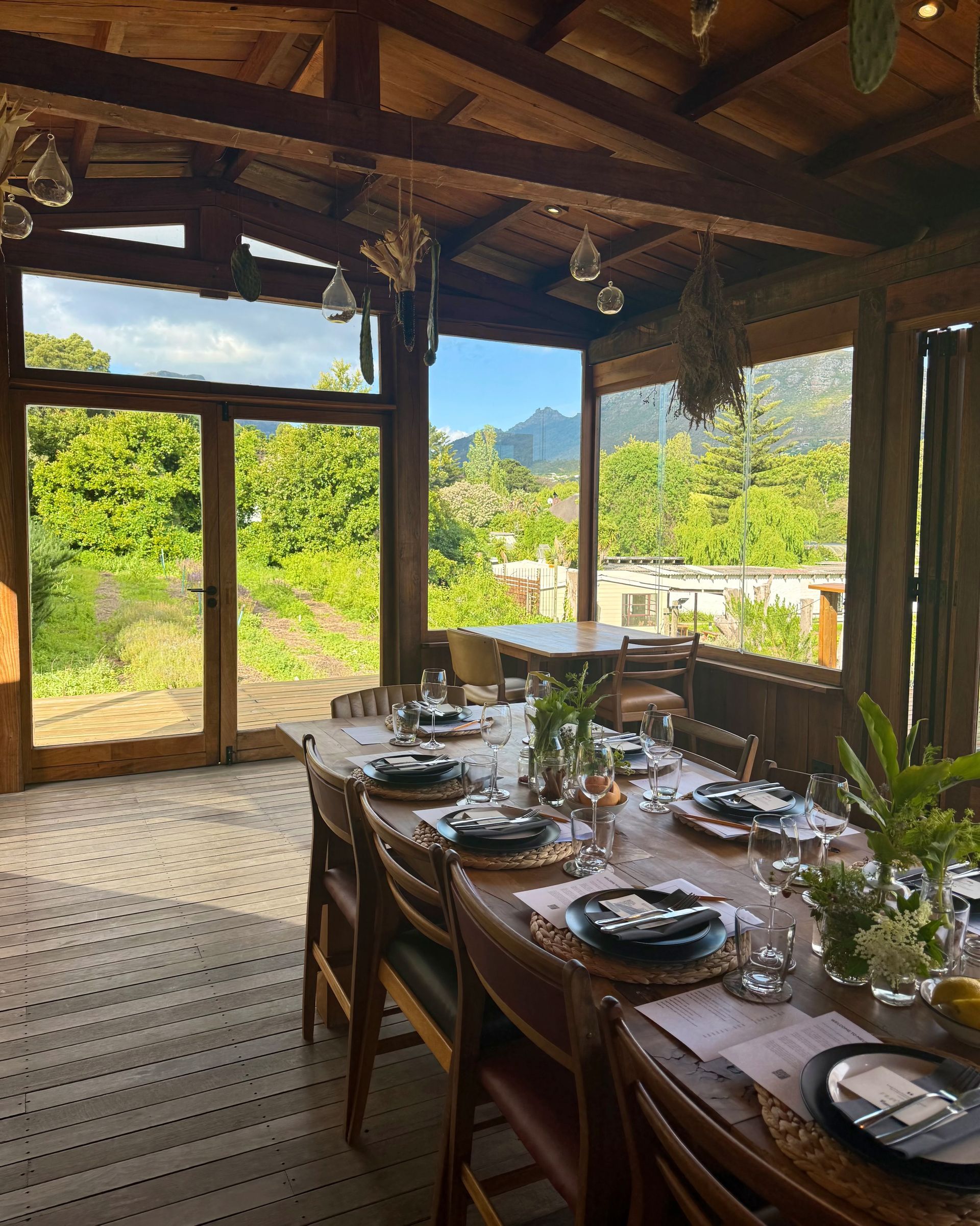 Wooden dining room with a long table set for a meal, overlooking a green landscape and mountains.
