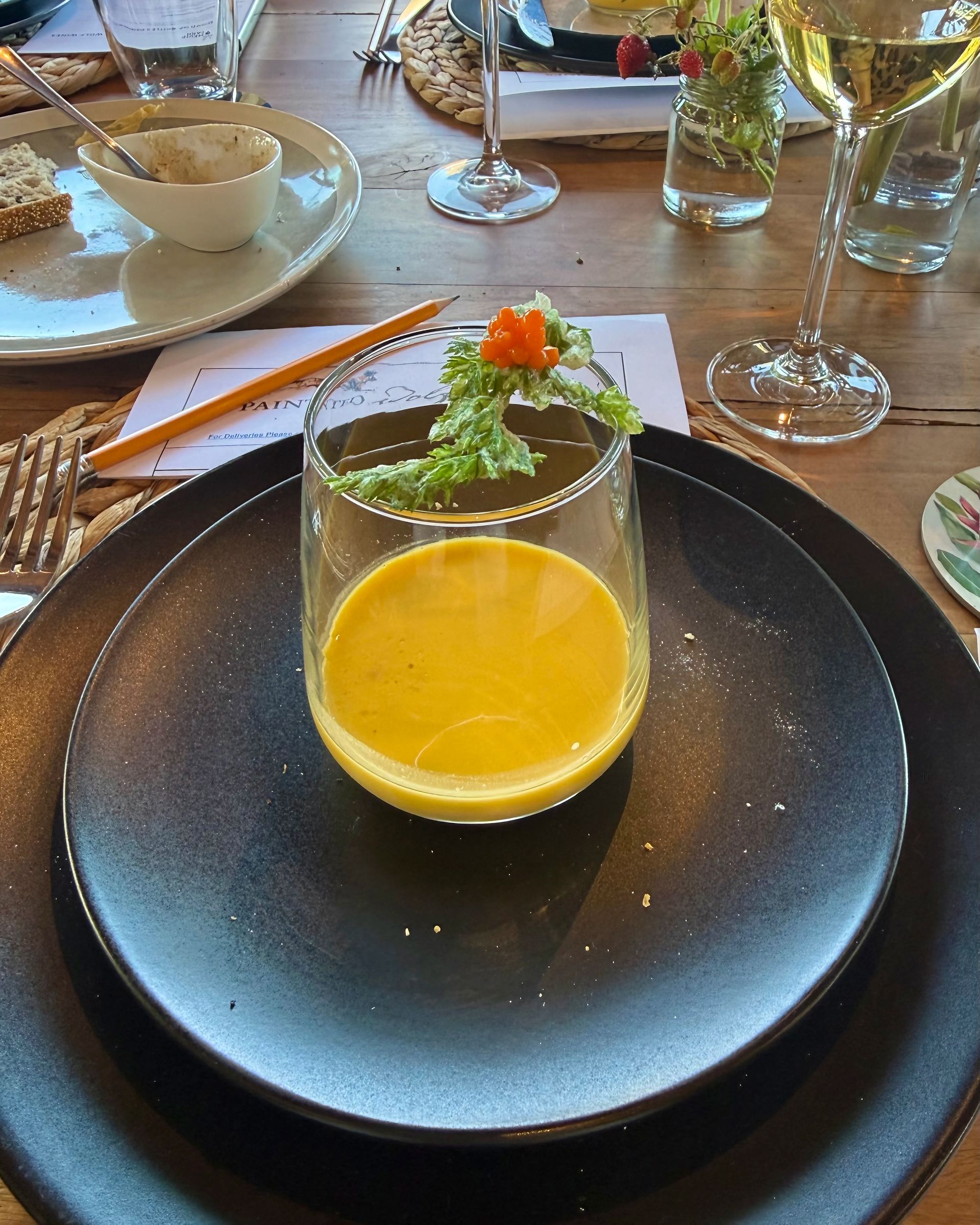 Yellow soup in a glass on a black plate, garnished with greens and pepper. Table setting.