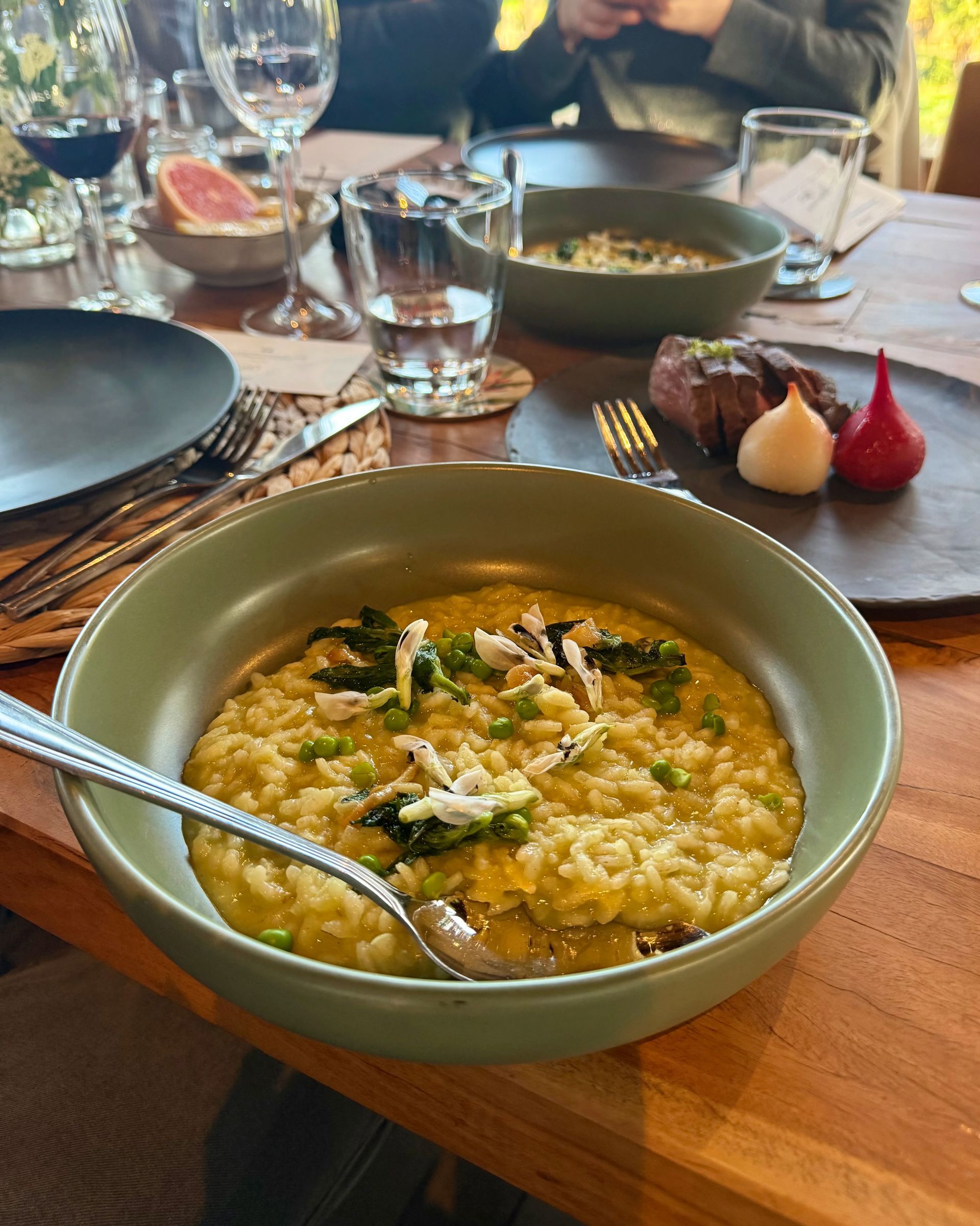 Risotto in green bowl, table setting with steak, drinks, and other dishes. Wooden table.